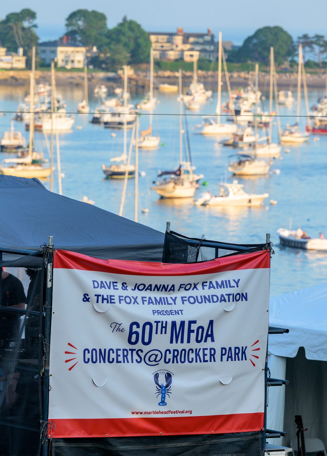 A banner announcing the 60th MFoA Concerts at Crocker Park hangs from the stage rigging above the harbor.