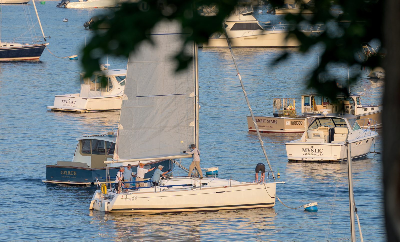 A sailboat crew gathers in the mainsail amid the moored boats of Marblehead Harbor as evening light settles over the water.