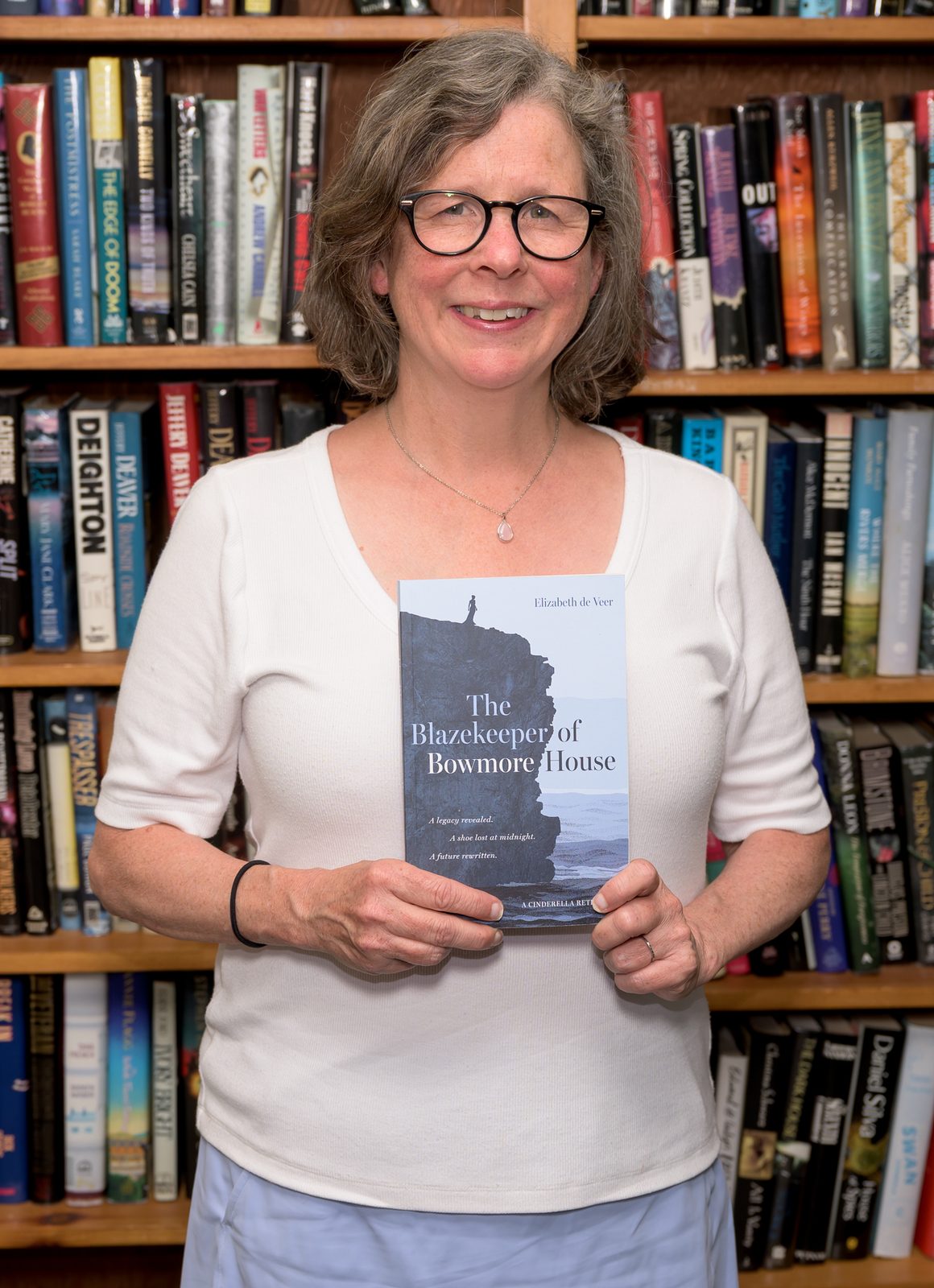 Elizabeth de Veer holds up a copy of her novel ‘The Blazekeeper of Bowmore House,’ a Cinderella retelling, before the used-book shelves.