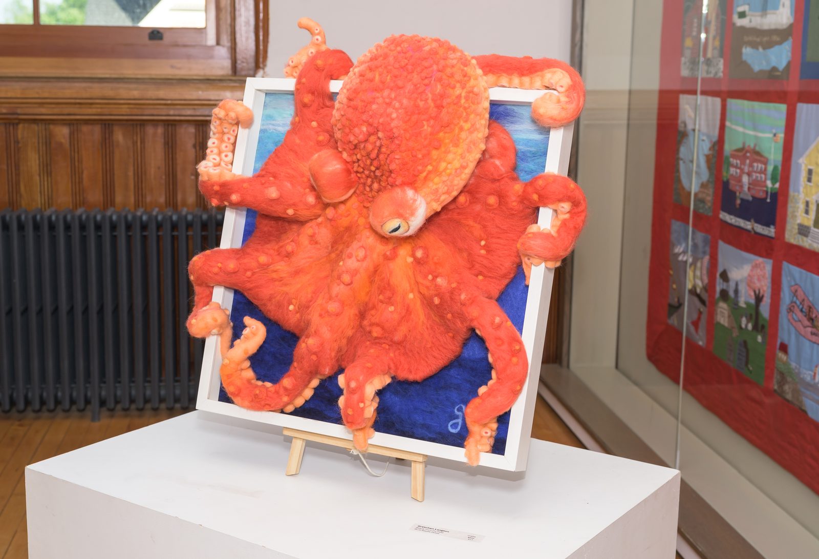 A needle-felted orange octopus climbs out of its frame in the gallery exhibition.