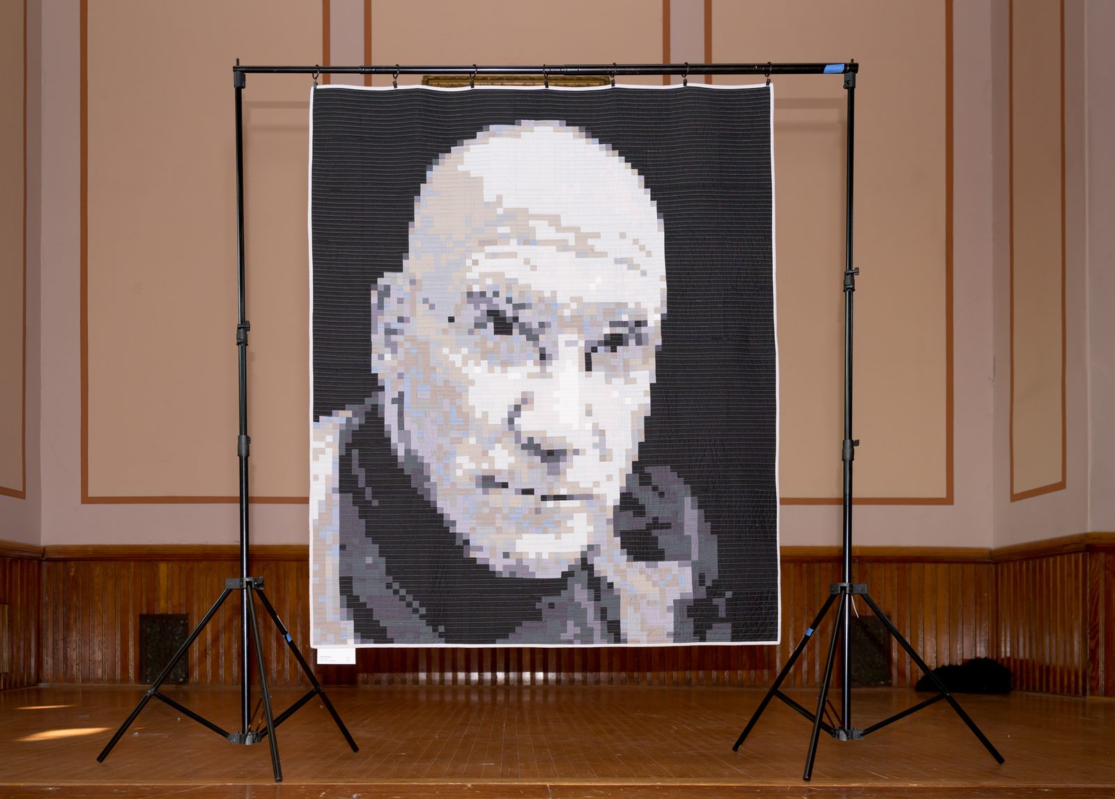 A wall-sized quilt renders a portrait in pixel-like fabric squares on a stage at the exhibit.