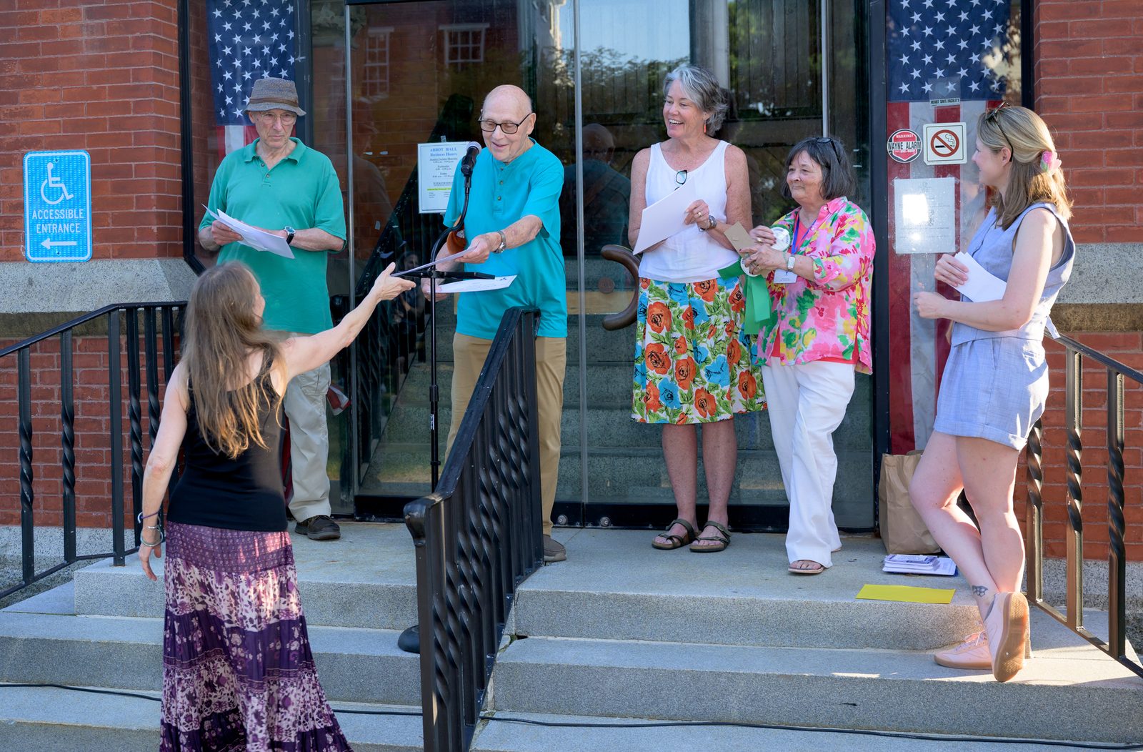 Clem Schoenebeck leans over the railing to hand down a certificate to a winner reaching up from the steps below, as Maile Black and, to her right, Margo Steiner look on smiling.