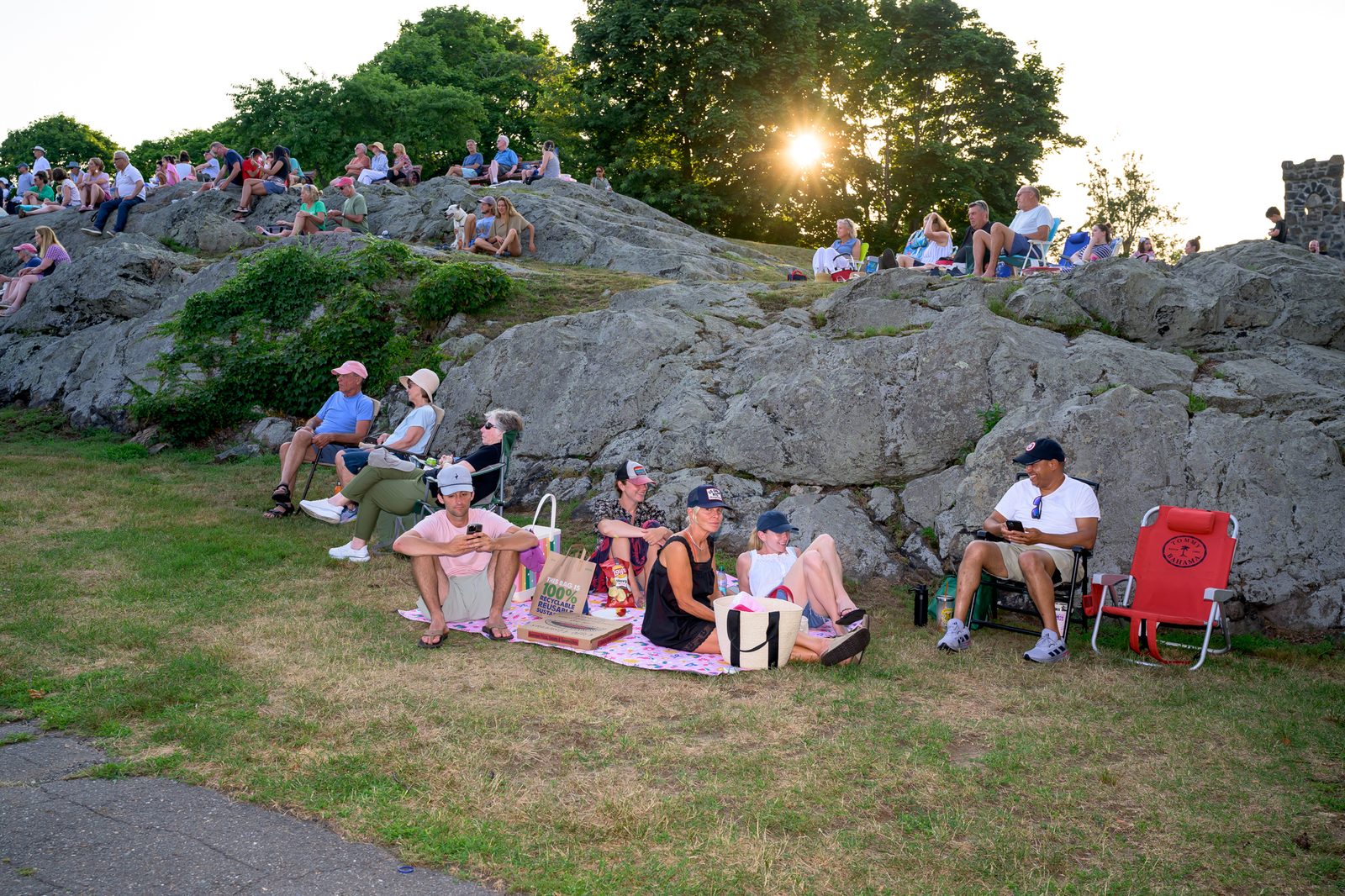 The evening sun breaks through the trees as concertgoers stake out spots on the rocks before a performance.