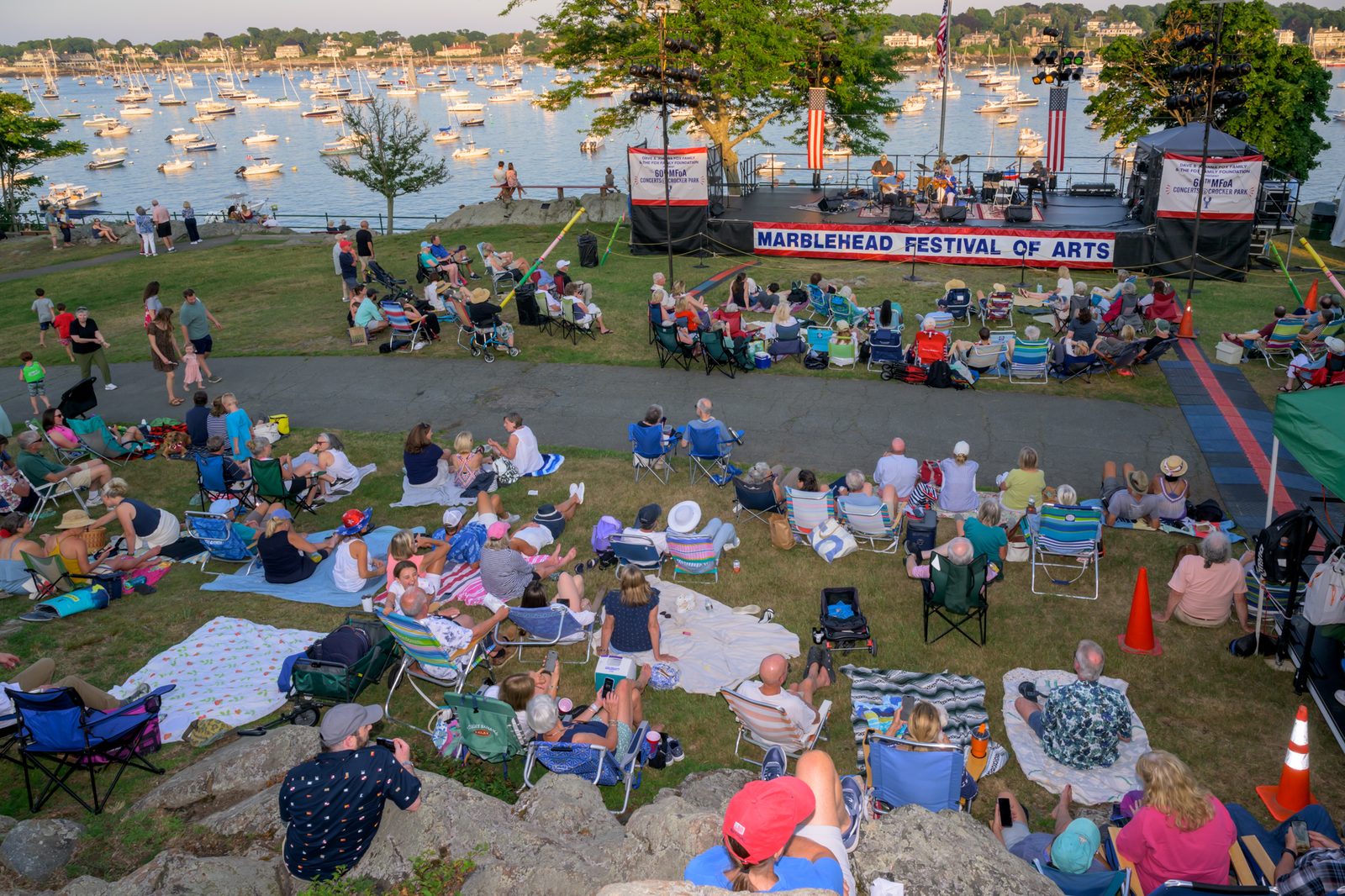 Concertgoers fill the lawn at Crocker Park as golden evening light falls over Marblehead Harbor during a 60th Marblehead Festival of Arts concert.