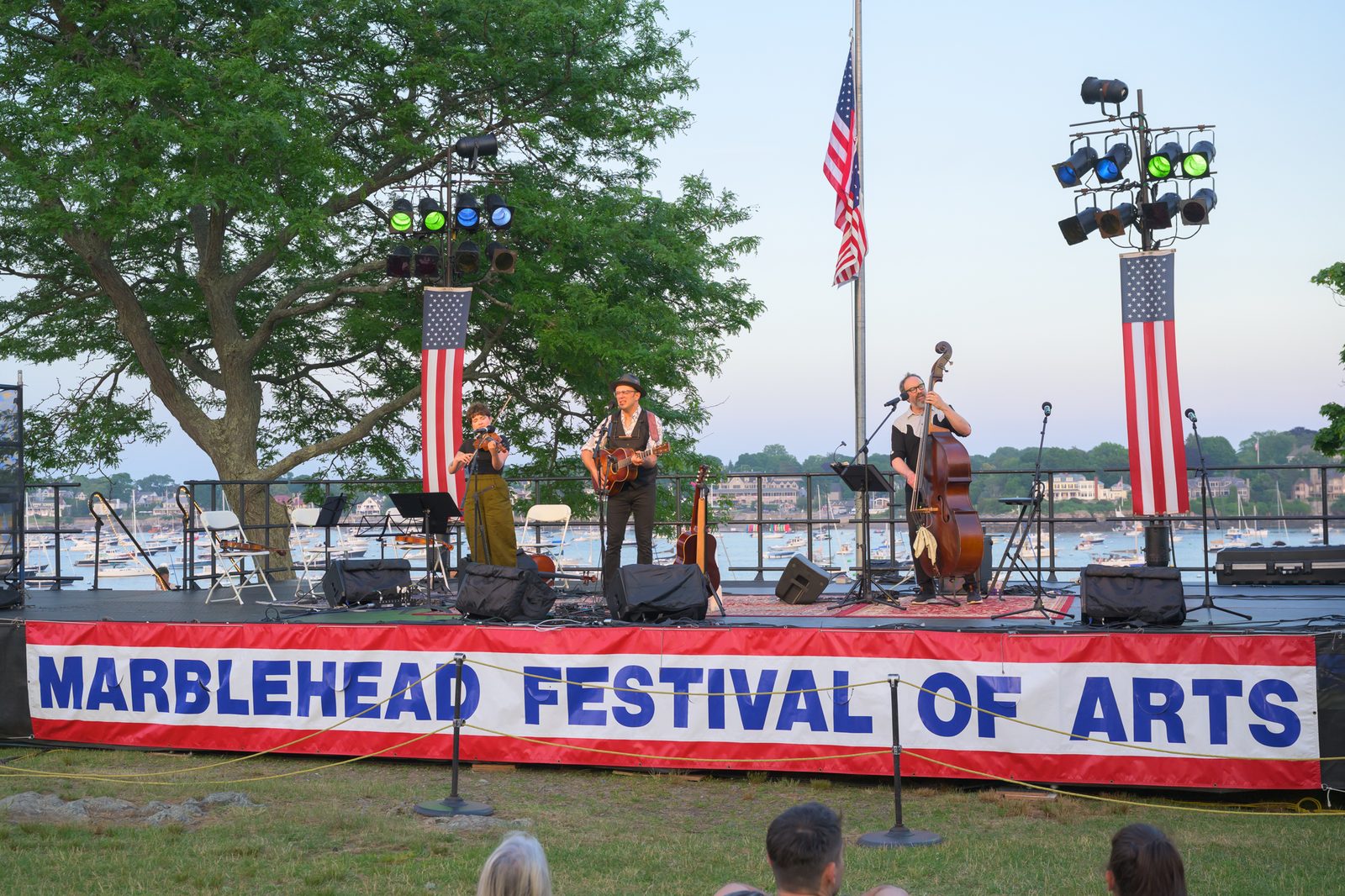 A folk trio plays fiddle, guitar and upright bass on the harborside main stage as twilight falls over the boat-filled harbor.