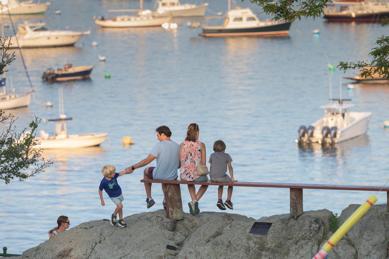 A young boy tugs at his father’s hand as a family takes in the view of the harbor from a rocky overlook.