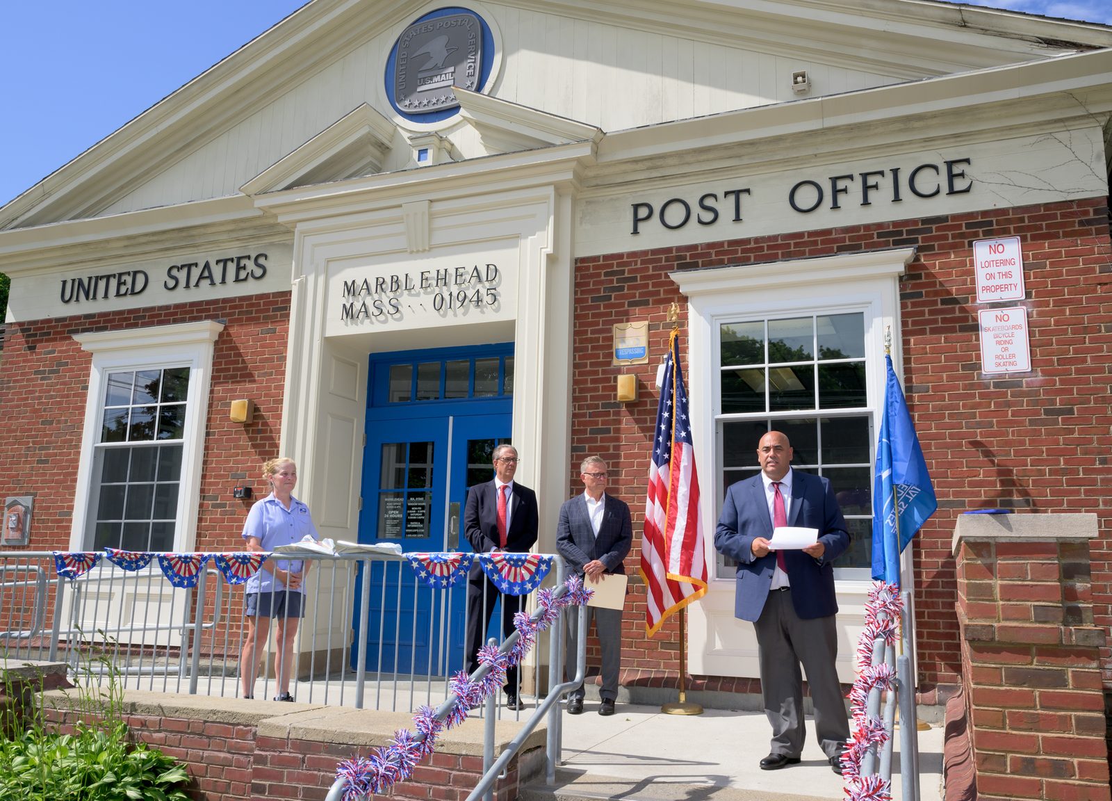 Postmaster Chris King reads from prepared remarks beneath the eagle seal as Select Board member Moses Grader and other officials look on.