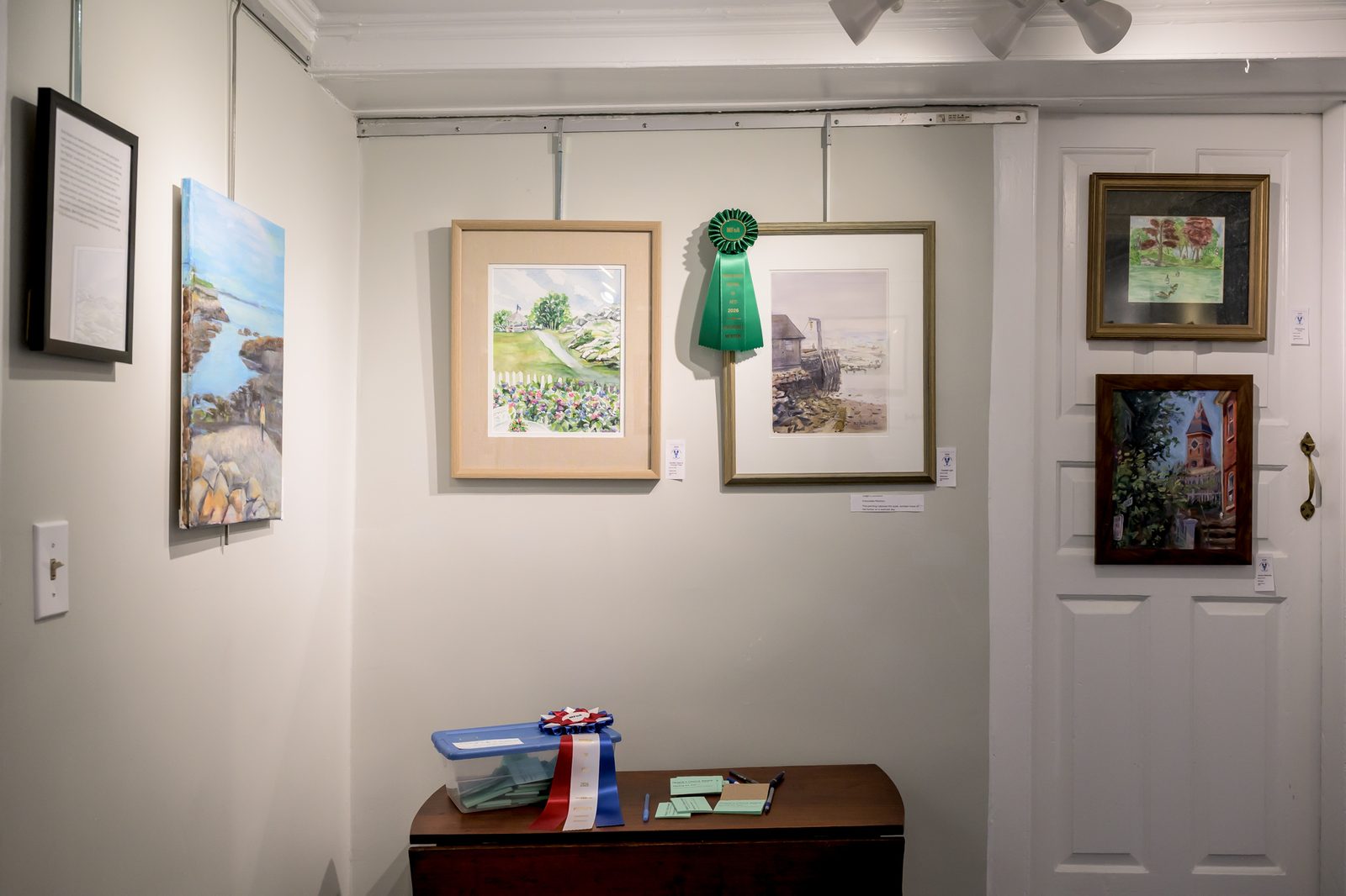An Honorable Mention ribbon hangs beside a harbor watercolor while People’s Choice ballots pile up at the Painting the Town exhibit.