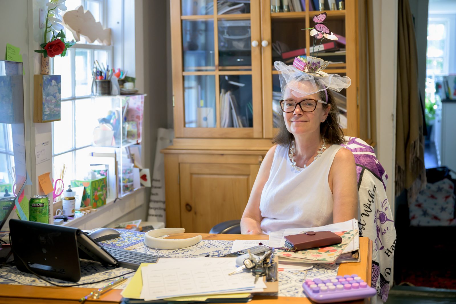 Marblehead Arts Association Executive Director Xhazzie Kindle, in a teacup-topped fascinator, smiles from behind a busy desk during festival week.