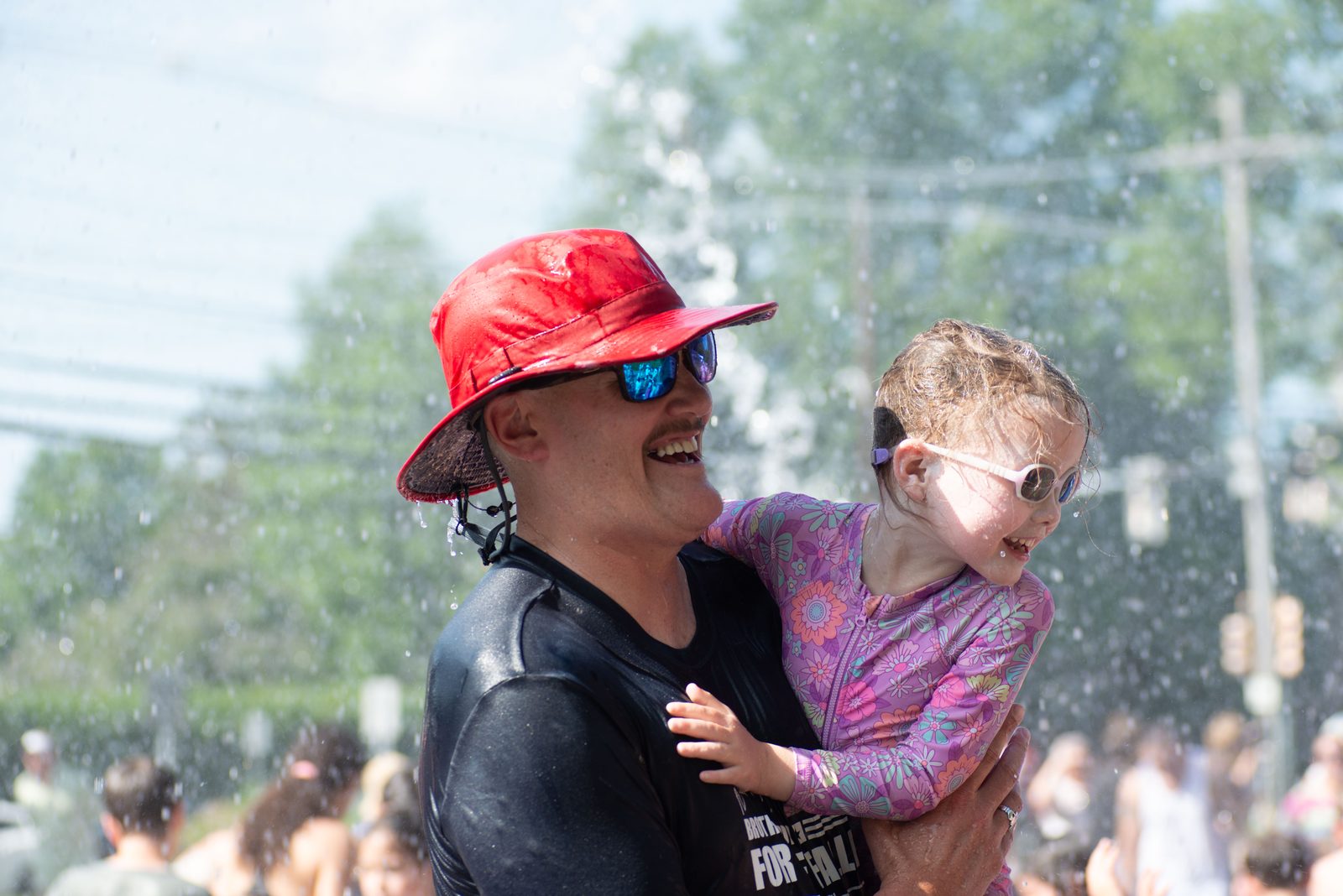 A man in a red bucket hat holds a small girl, both soaked and grinning.