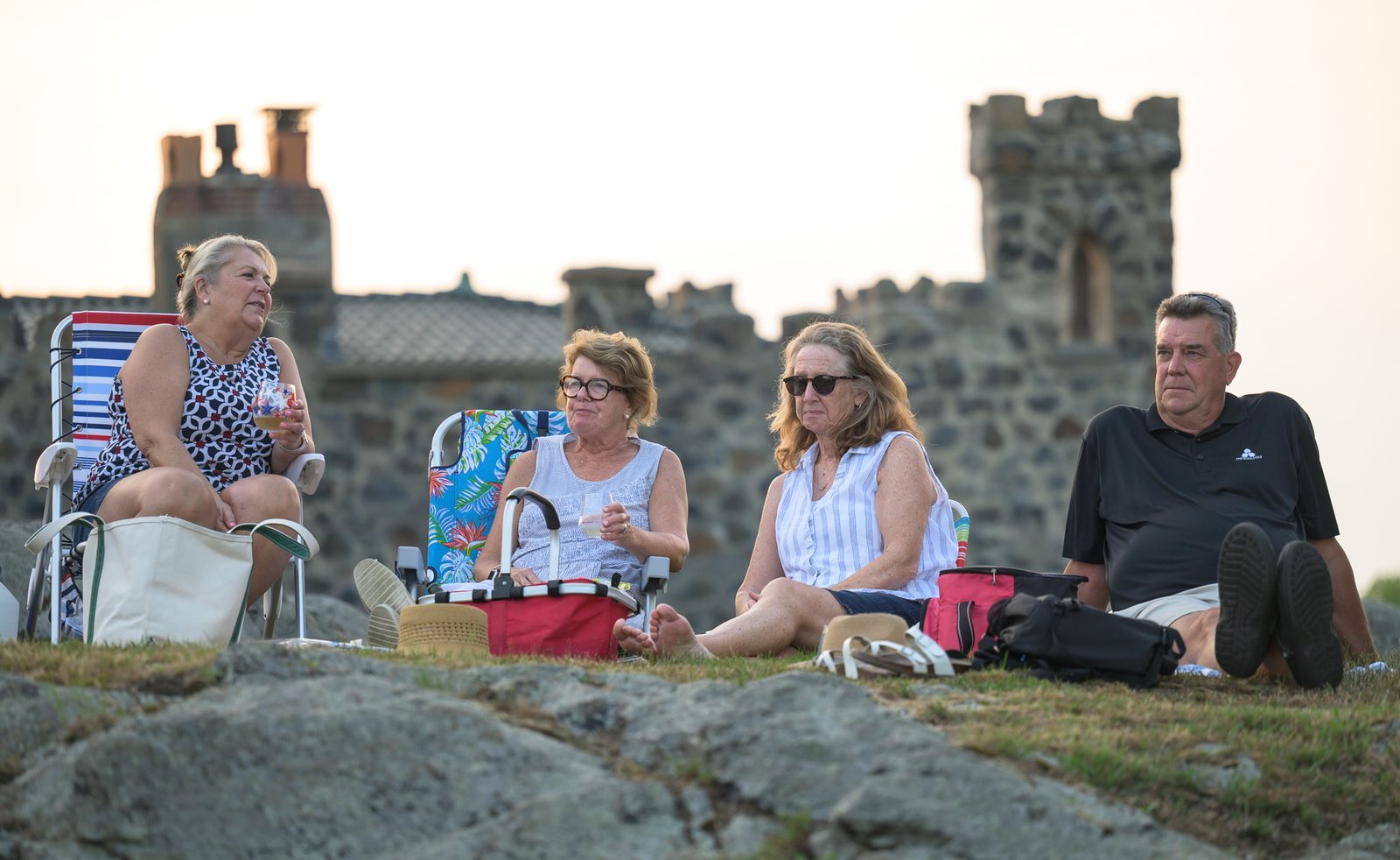 Four listeners tip their beach chairs toward the water below a castellated stone tower as the evening light softens.
