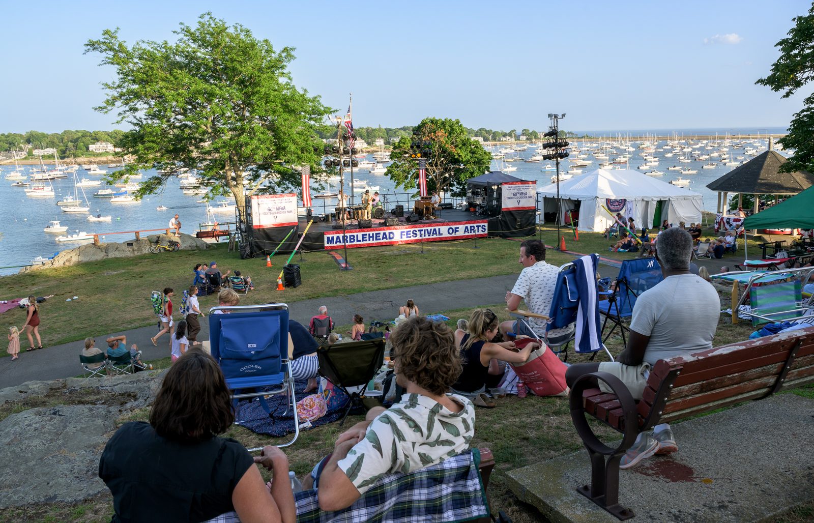 Concertgoers settle into beach chairs on the slope at Crocker Park as the band plays beneath the Marblehead Festival of Arts banner, the harbor full of moored boats beyond.