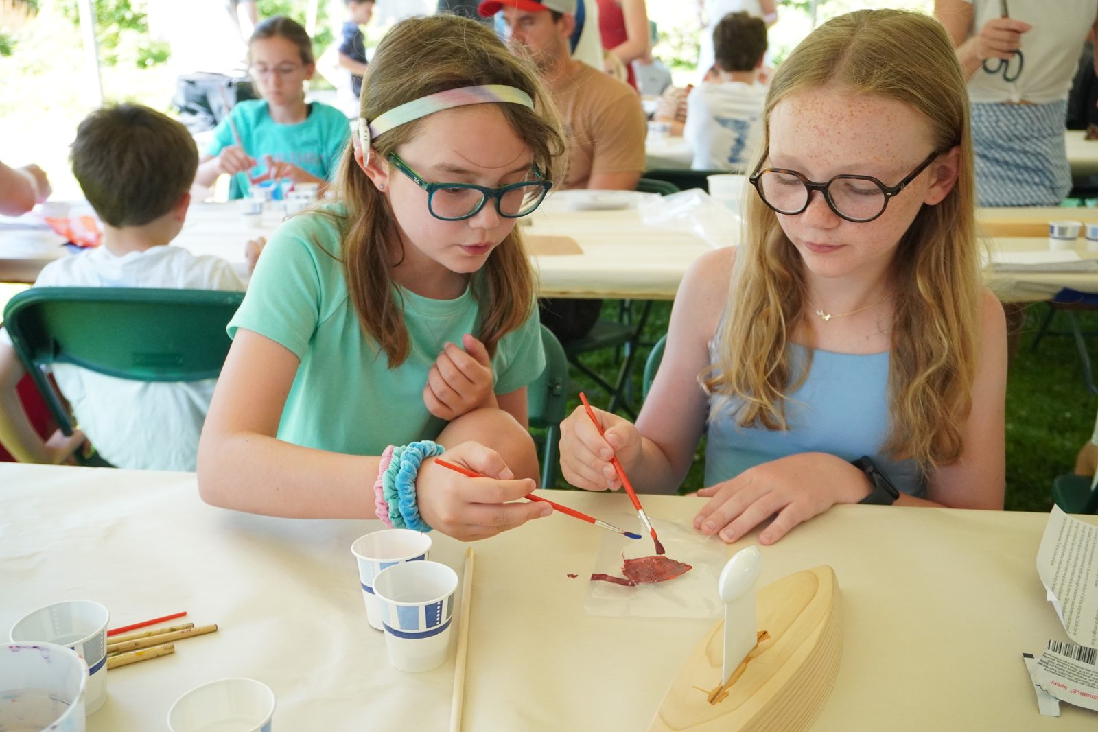 Two friends paint their wooden boats side by side under the tent.