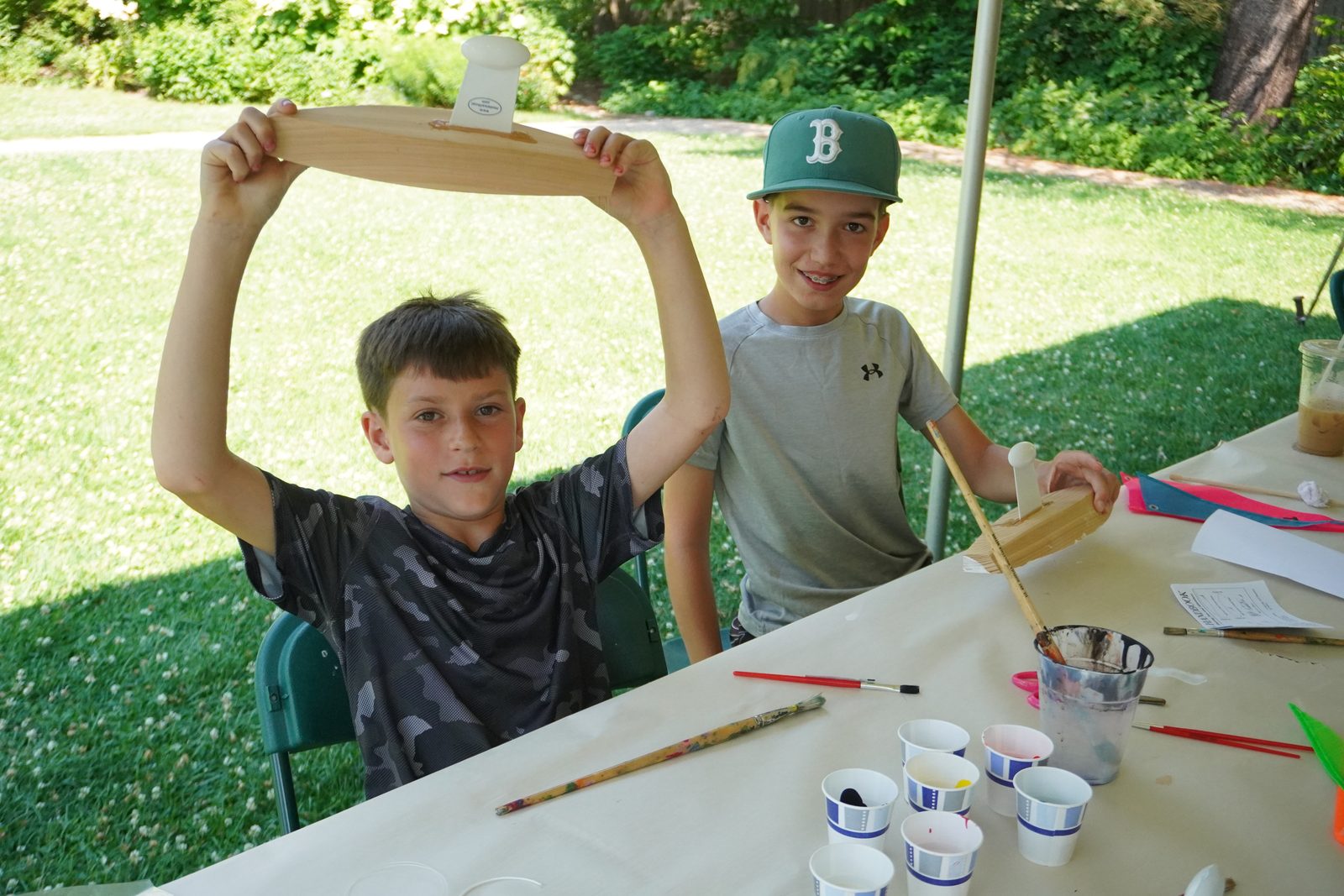 A boy holds up his finished hull like a trophy as a friend looks on.