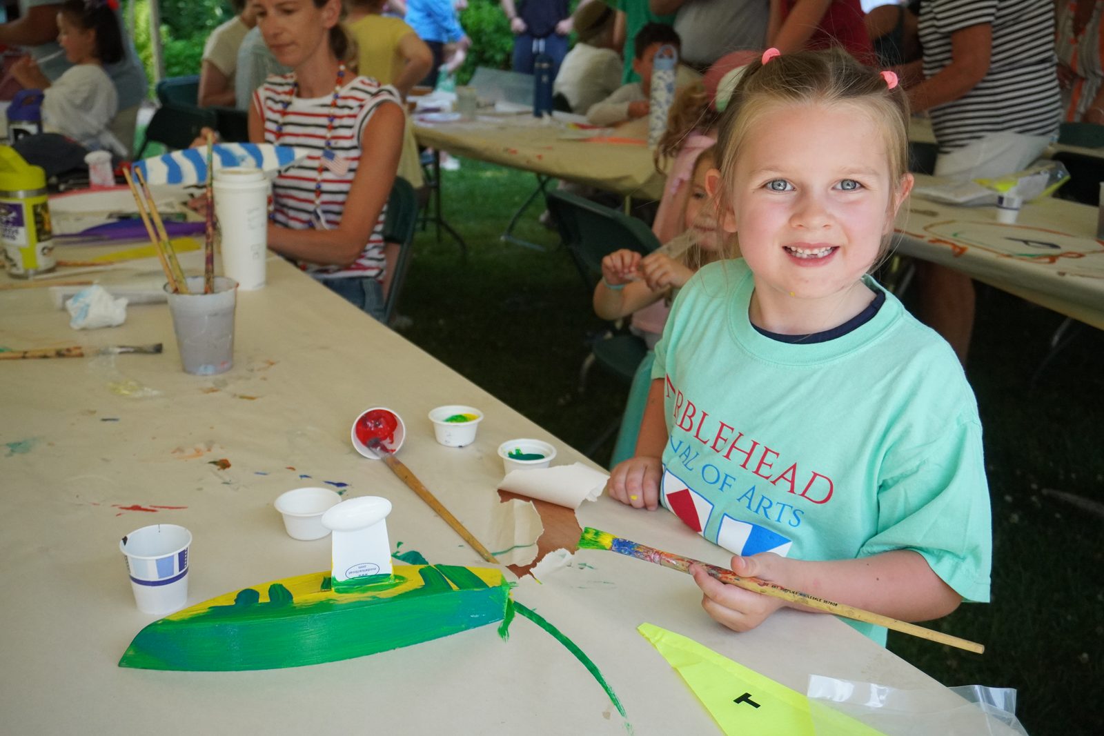 A girl in a Festival of Arts shirt beams over her green-and-yellow boat.