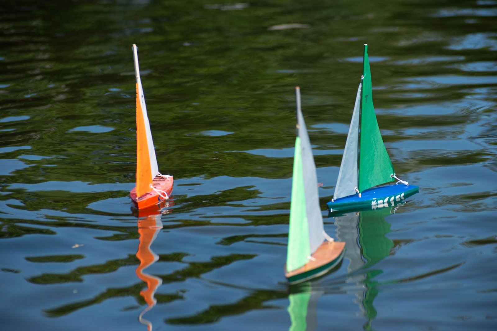 Three finished sloops — orange, white and green — ride the pond on a light breeze.