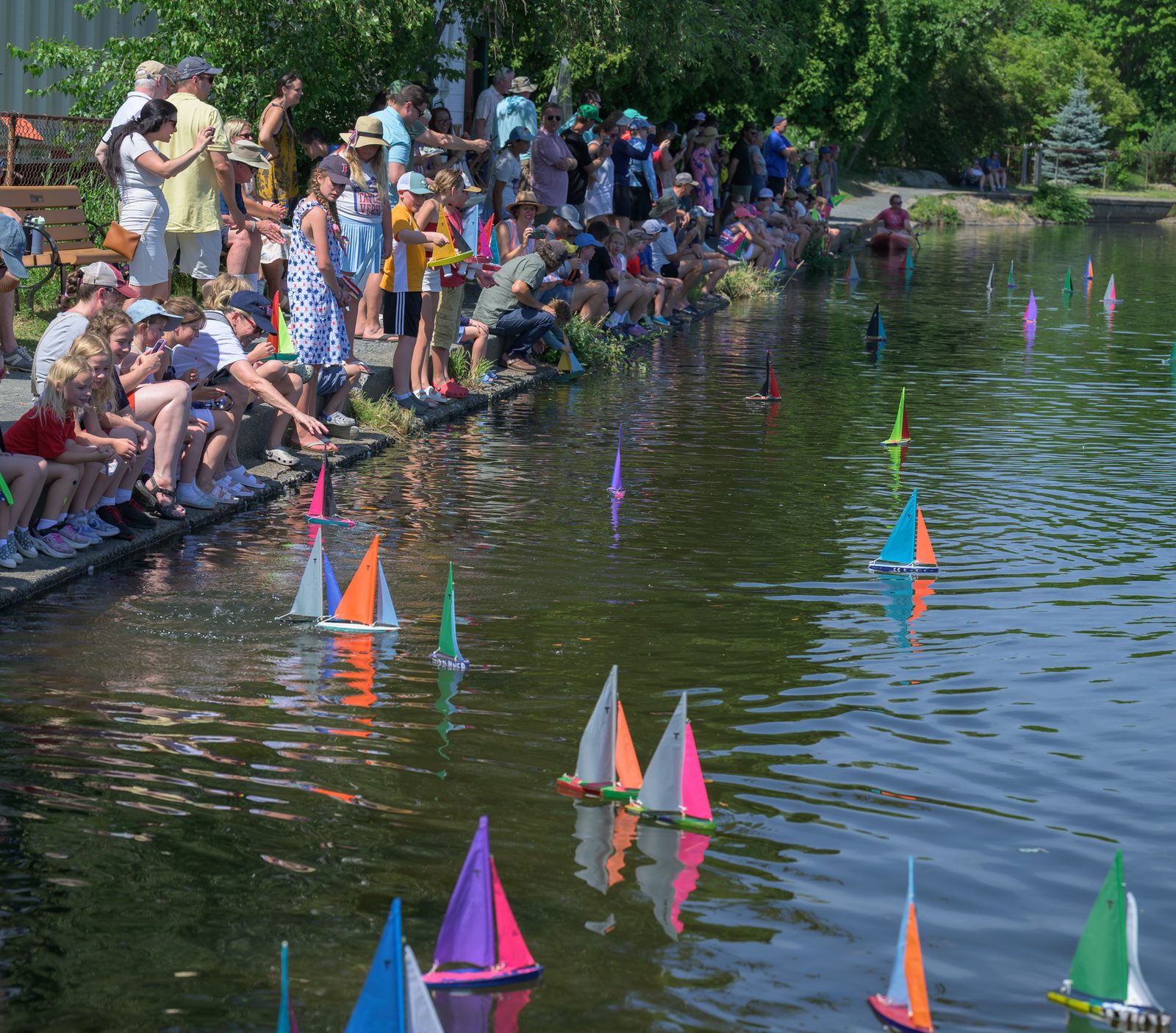 A fleet of model boats scatters across the pond as families watch from the far bank.