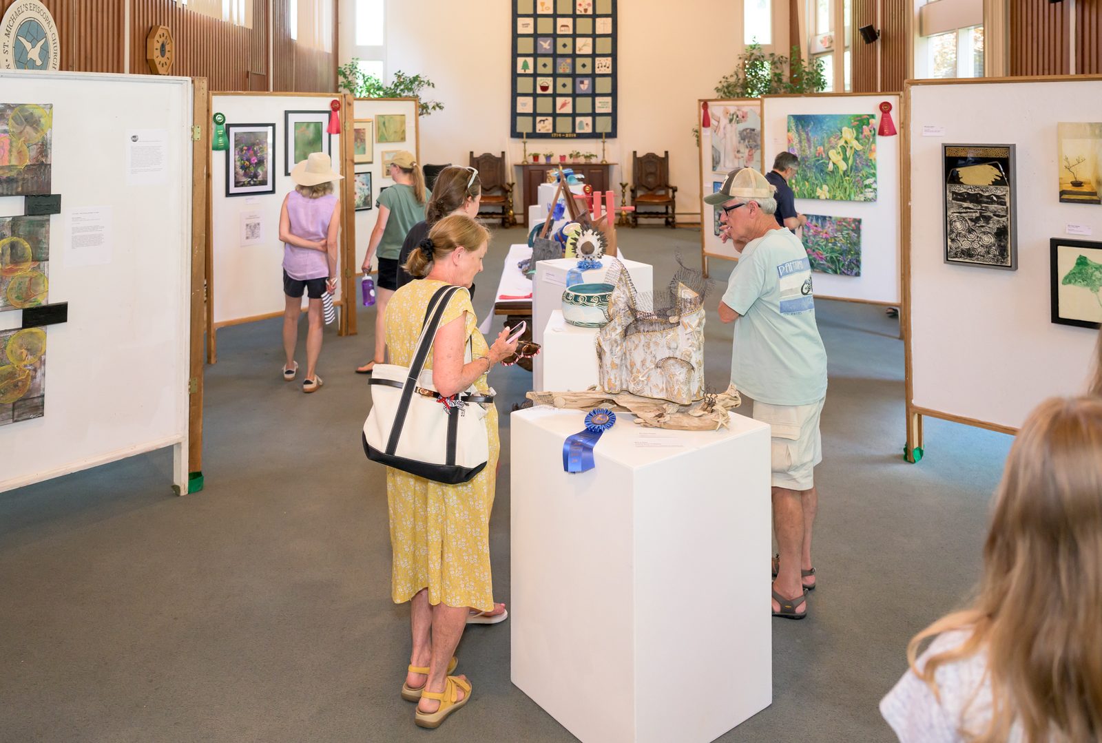 Inside St. Michael’s Episcopal Church, visitors move among the festival’s juried exhibits, where a blue ribbon marks a first-place work of driftwood and shell.