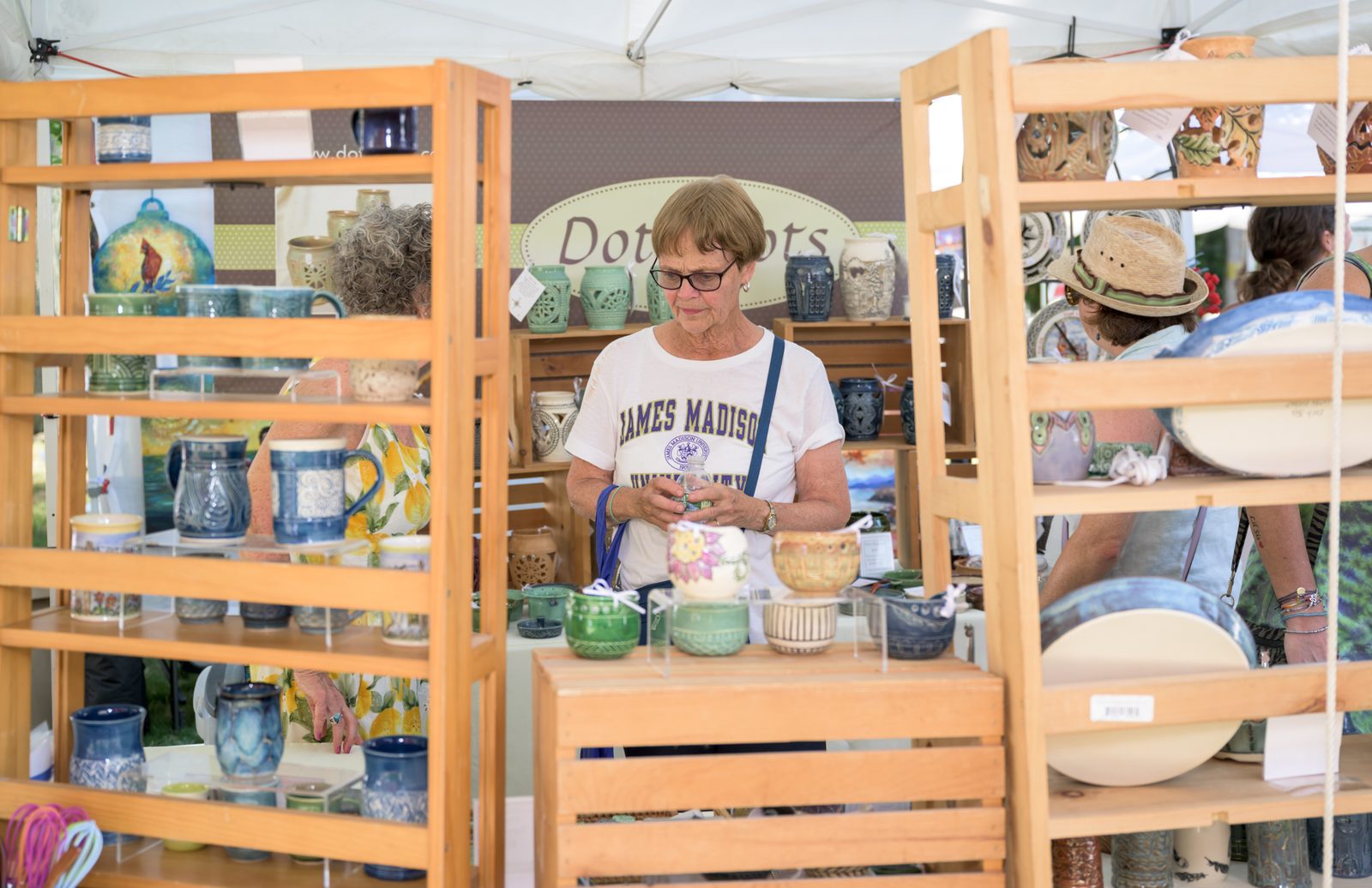 A shopper considers a piece at a pottery booth, its shelves lined with glazed mugs, vases and bowls.