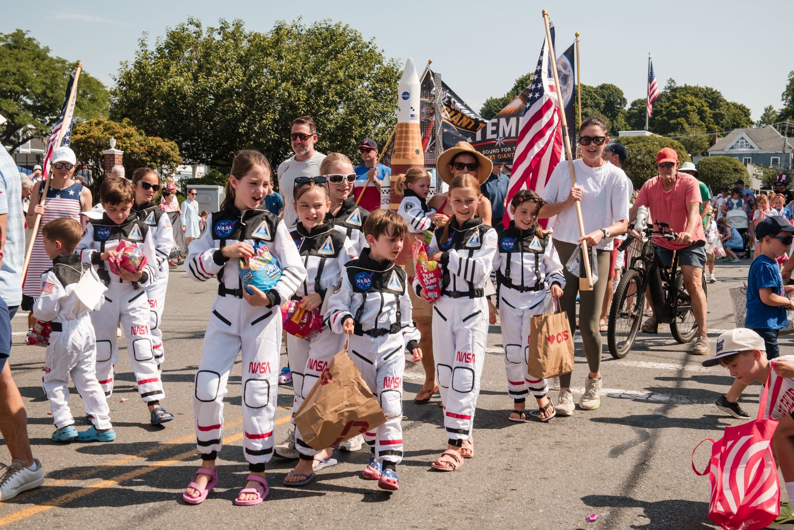 Children in NASA flight suits march behind a homemade rocket, one carrying an American flag.