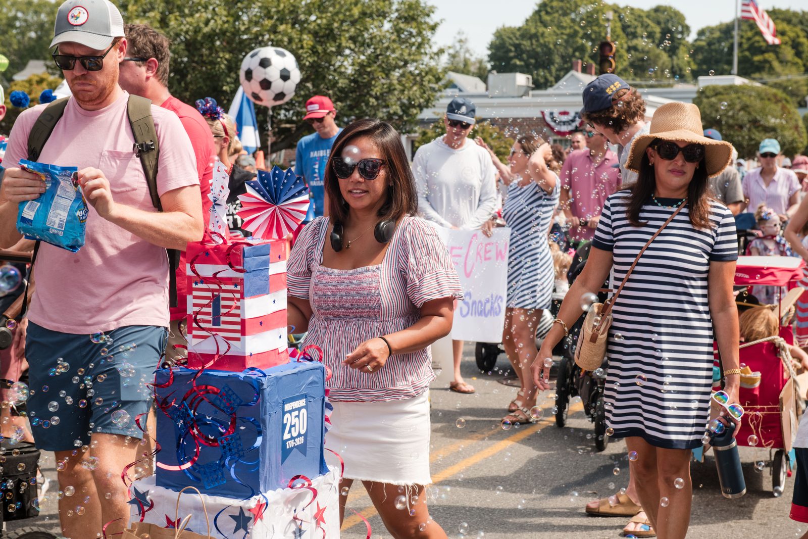 A stack of gift-wrapped boxes on a wagon spells out ‘Independence 250, 1776–2026’ amid a cloud of bubbles.
