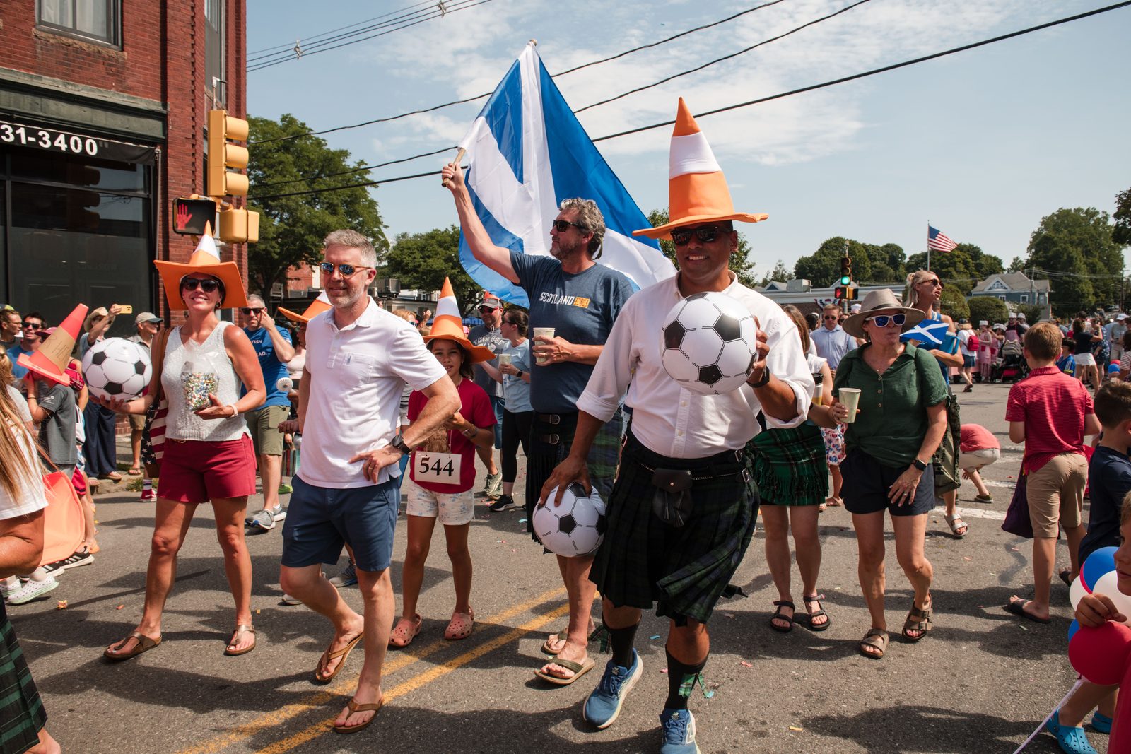 A soccer contingent in Scotland flags and traffic-cone hats crosses the intersection by the Chamber of Commerce information booth.