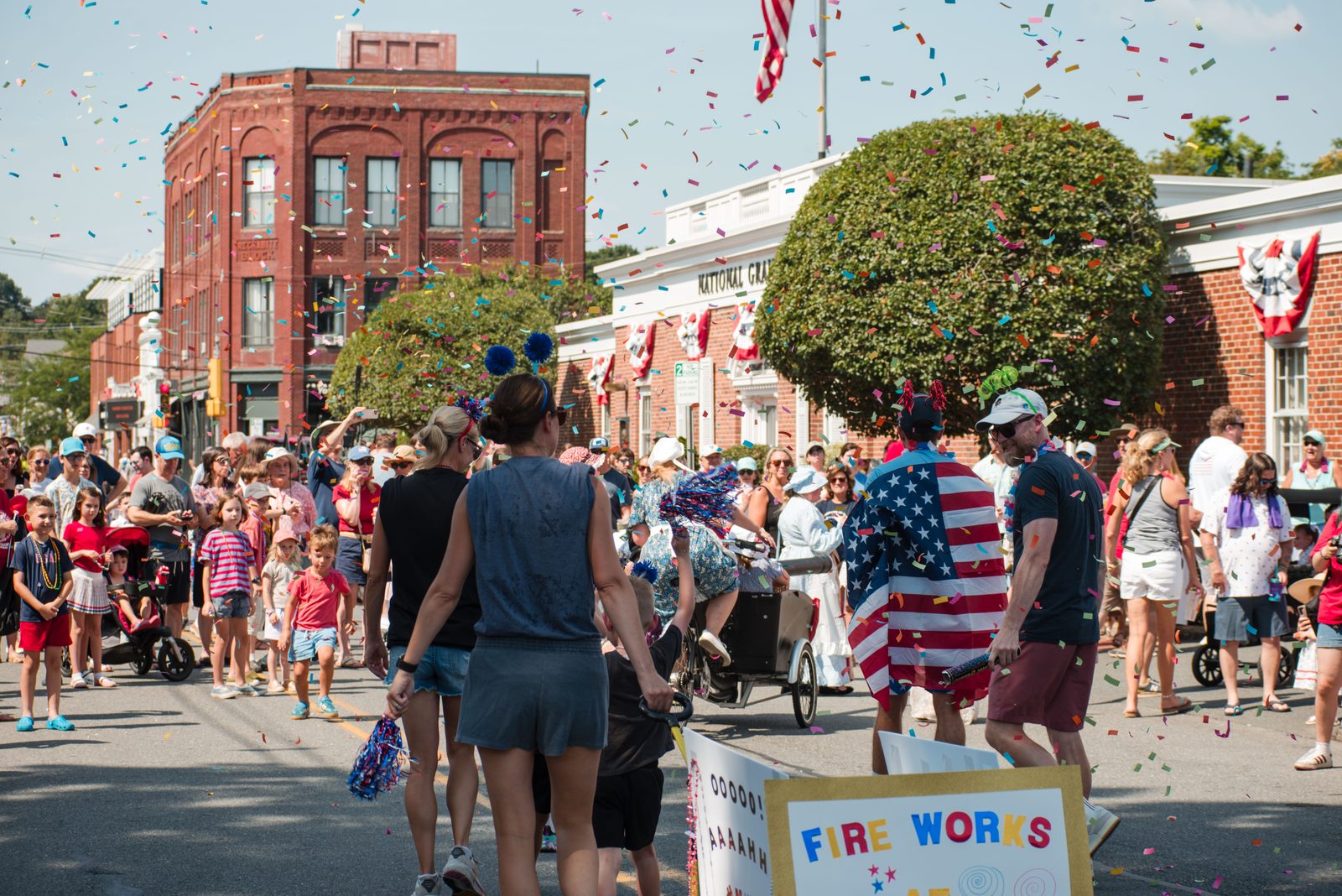 Confetti rains over the street as a marcher in a flag cape passes a child’s hand-lettered ‘Fire Works’ sign.