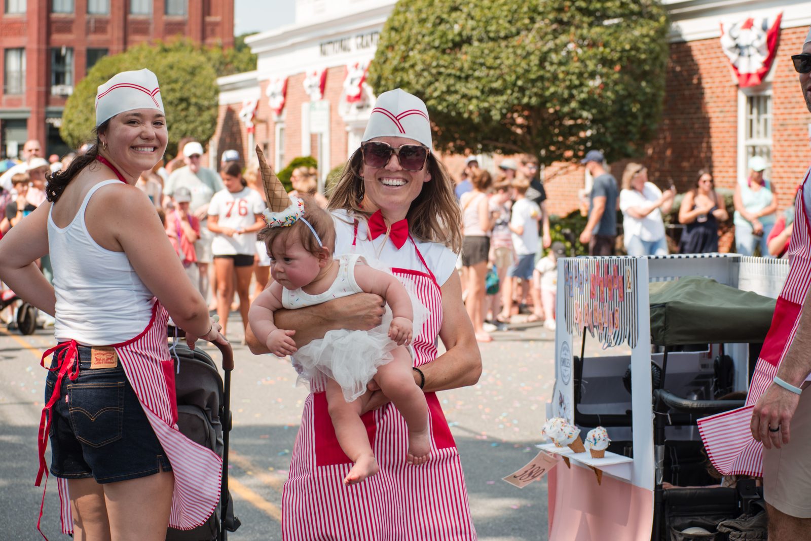 Marchers in striped soda-jerk aprons push an ice-cream stand, one carrying a baby in an ice-cream-cone hat.