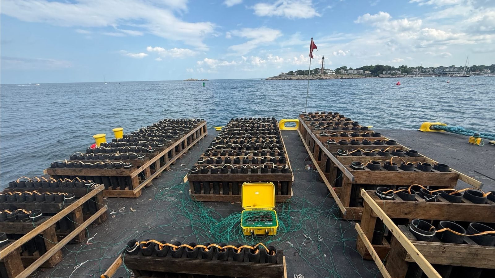 Racks of loaded mortar tubes cover the fireworks barge in the hours before the show, wired to the yellow electronic firing controller at center, with Marblehead Light on the point beyond.