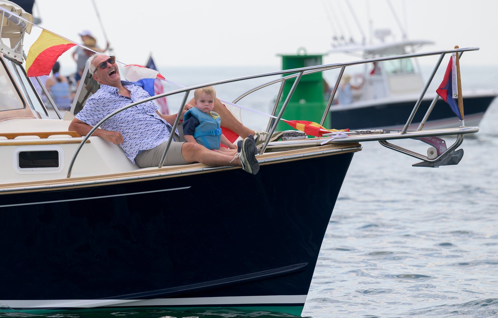 An older man and a small child laugh together on the bow of a flag-decked boat.