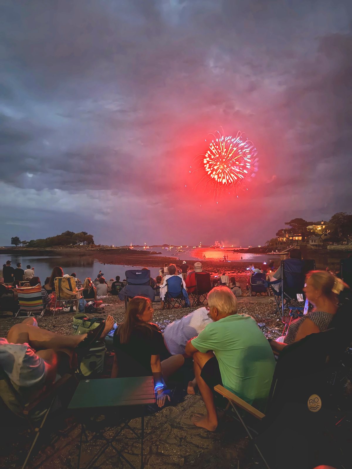 A red burst opens beneath low storm clouds at dusk, the harbor still faintly blue behind the crowd.