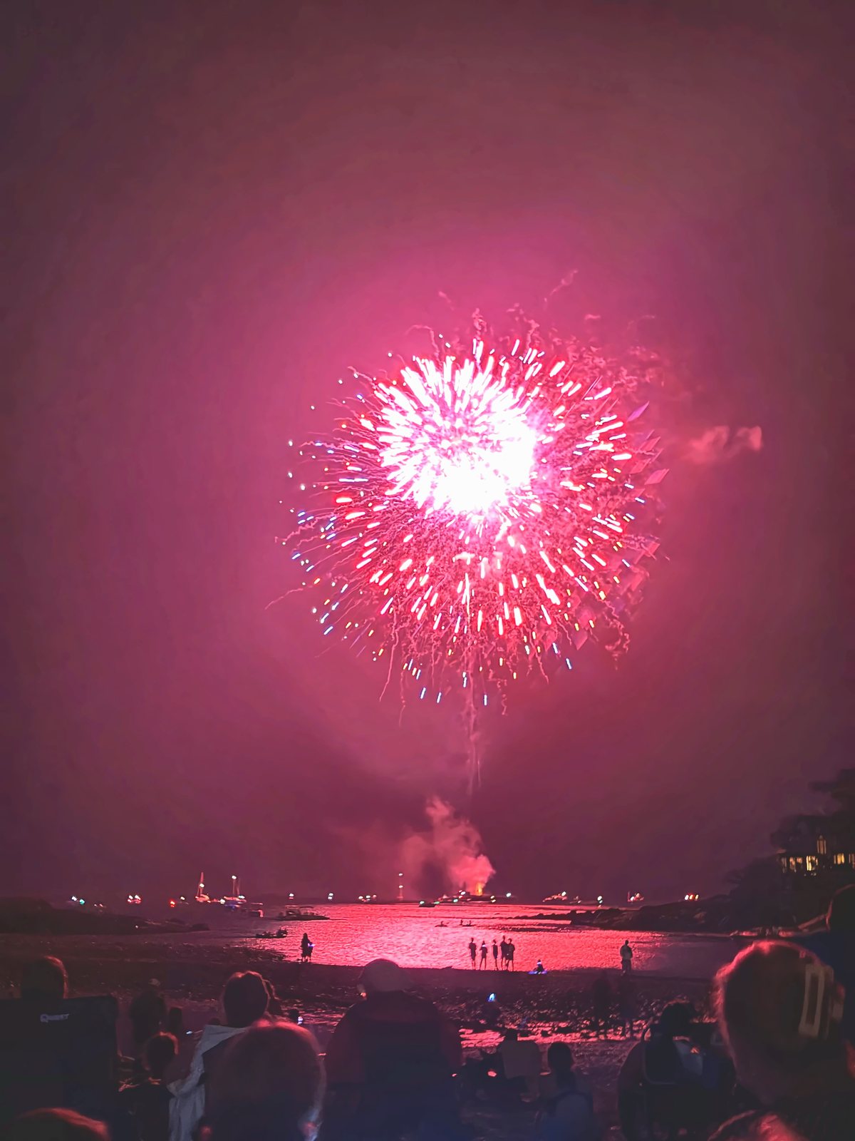 A tall gold burst rises over the harbor as spectators watch from the beach below.