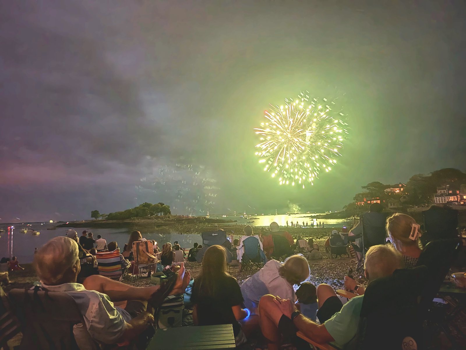 A green burst lights the sky and the water as the crowd watches from beach chairs along the shore.