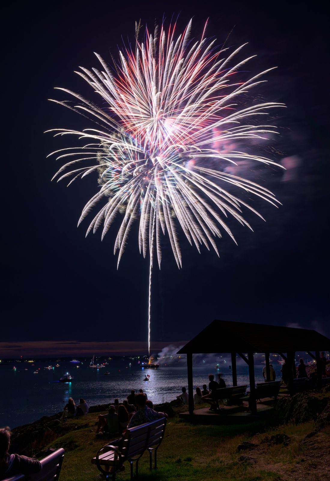 A single burst hangs over the harbor and the moored boats, the barge smoking below, as spectators watch from the shore.