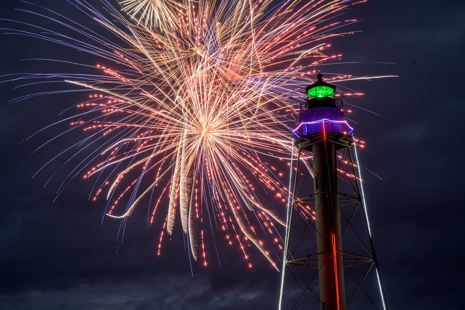 A burst opens beside Marblehead Light, its lantern lit green and the tower strung with color, over the harbor on the Fourth.
