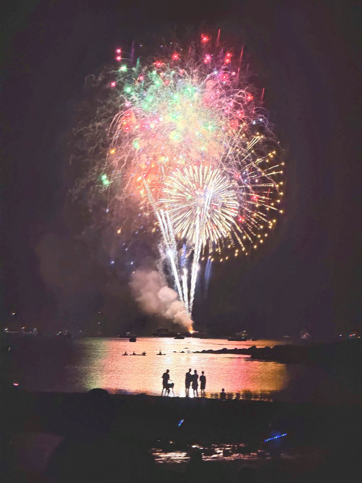 A wide multicolored burst fills the sky over silhouetted watchers at the water’s edge.