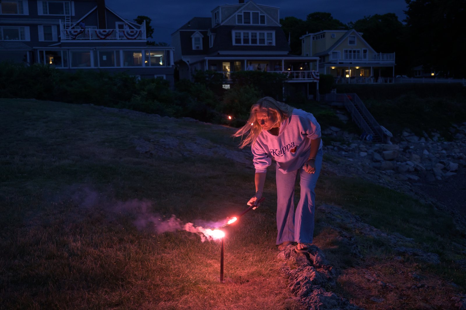 A flare catches and smokes as it is lit along the shore at dusk, houses glowing behind.