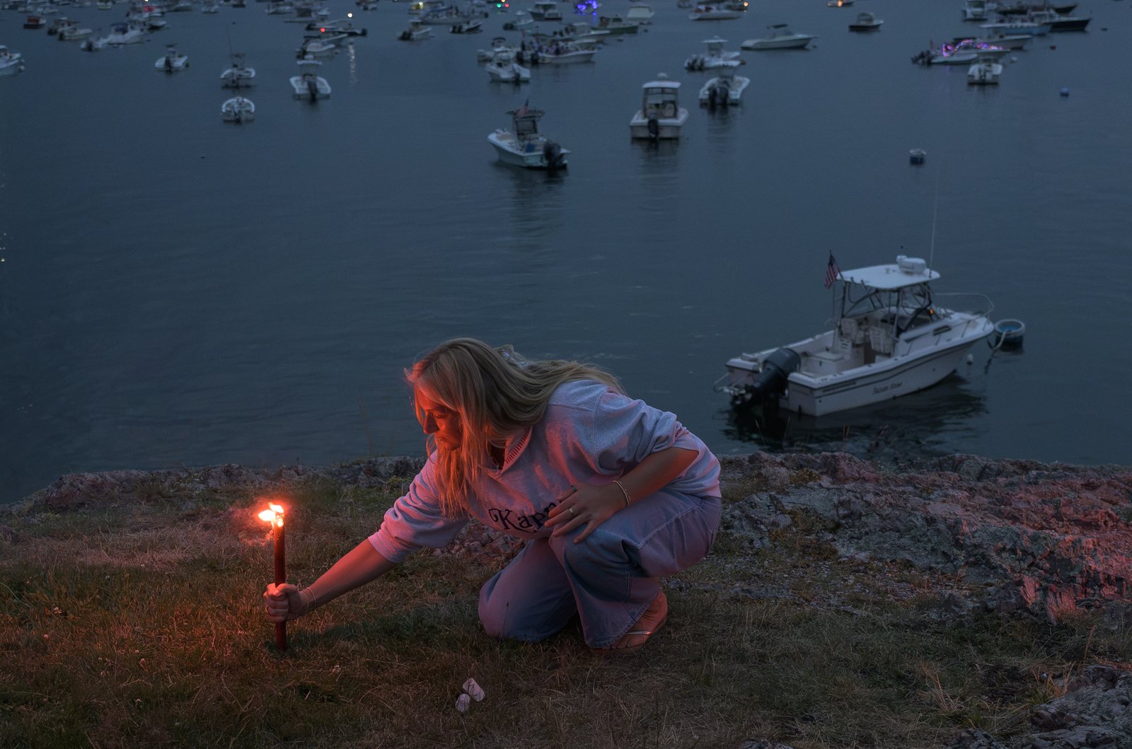 A woman kneels to light a red flare on the rocks above the harbor as boats lie at their moorings below.